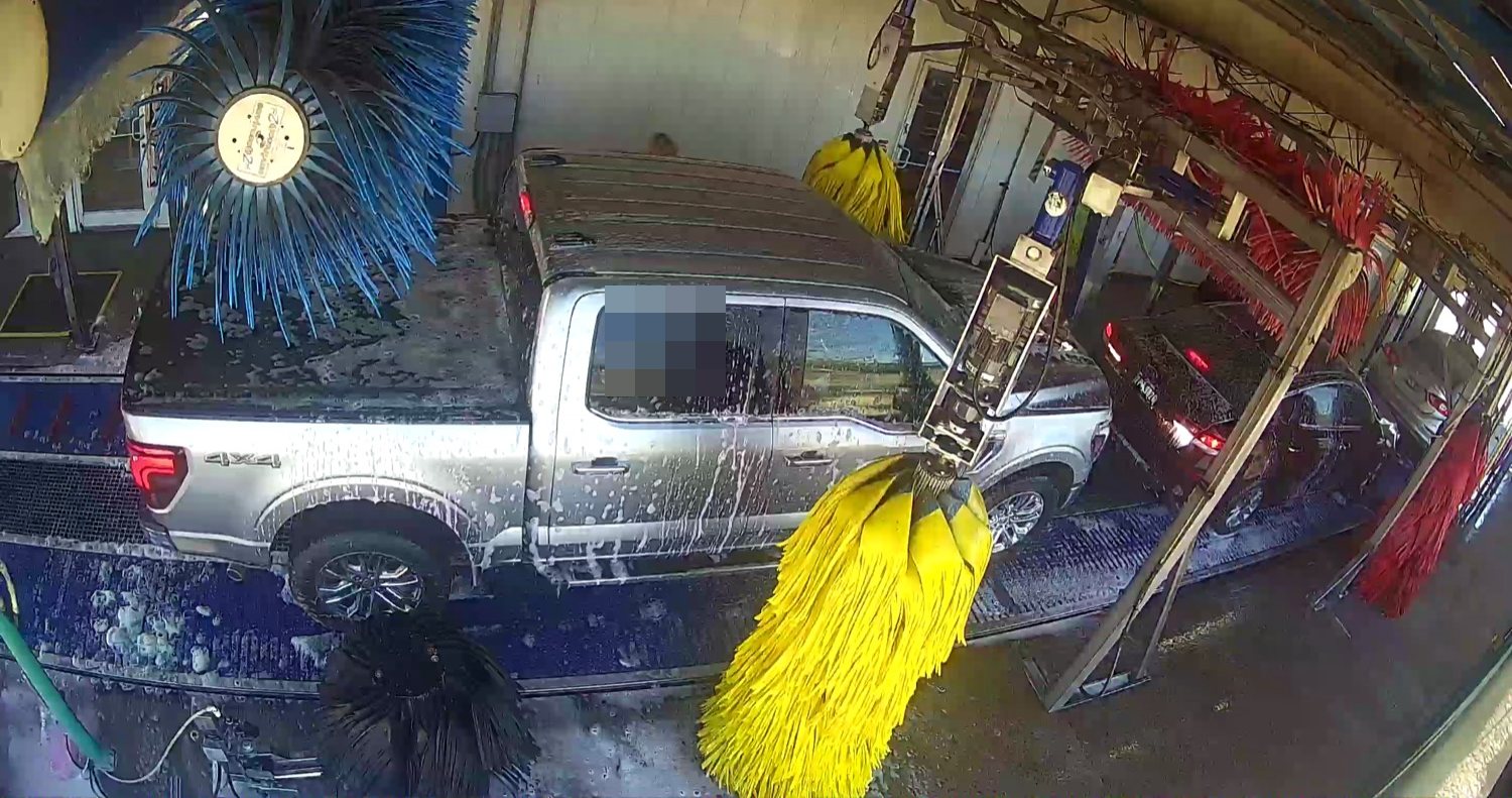 Wash-tunnel camera frame with a pickup partly crossed by brush equipment and a support assembly.